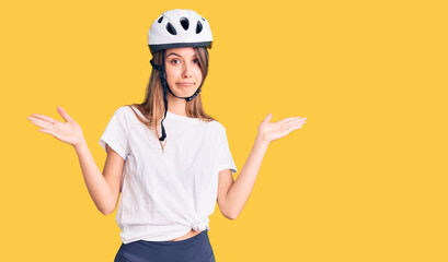 Young beautiful girl wearing bike helmet clueless and confused expression with arms and hands raised. doubt concept.