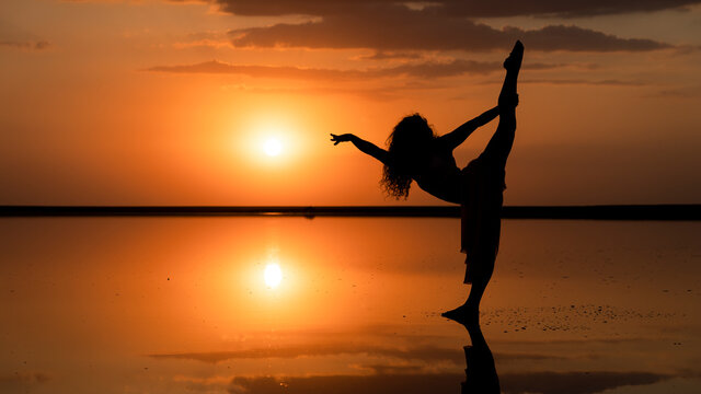 silhouette of a flexible girl performing split on the background of a golden sunset mirrored reflection in the water