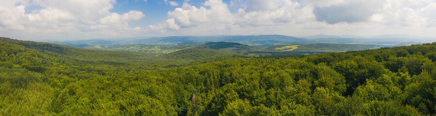 Vast summer scenery with green forested hills from aerial perspective. Wide panoramic composition of untouched nature sunlit in the morning. Blue sky with clouds above landscape.