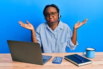 Young african woman wearing call center agent headset clueless and confused with open arms, no idea and doubtful face.