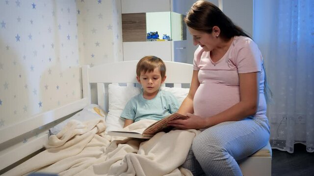 Beautiful pregnant mother readng story book to her older son lying in bed at night before going to sleep. Parents reading to children