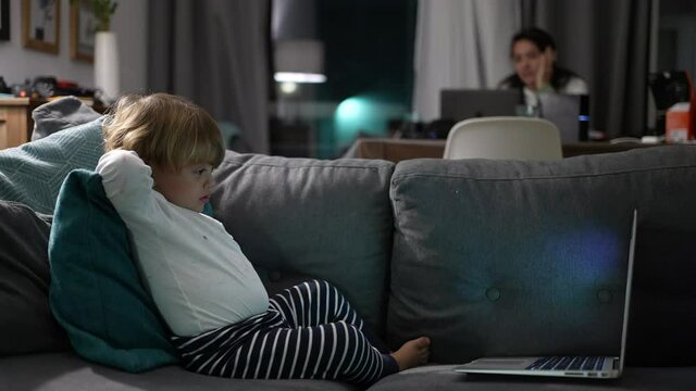 Child watching computer laptop screen while mother works at night