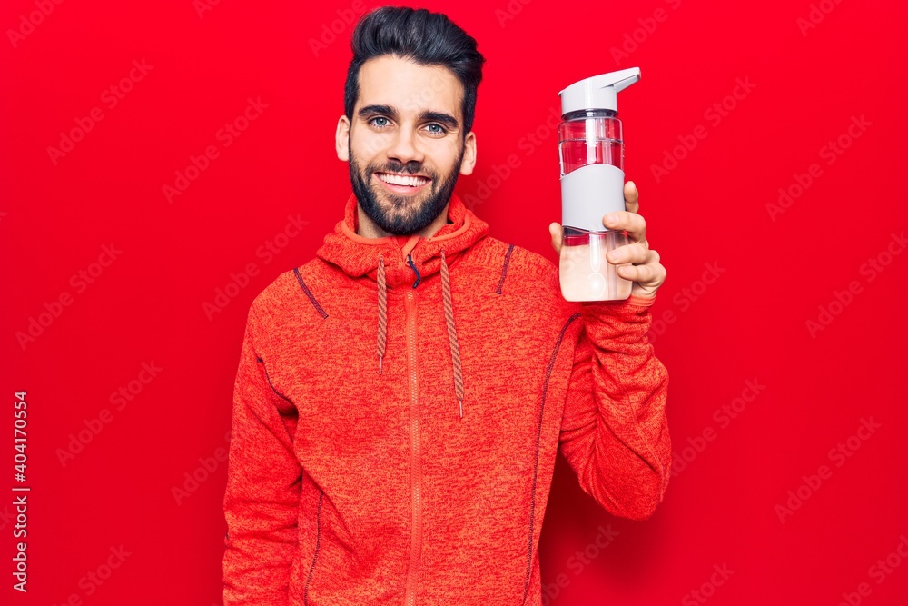 Wall mural young handsome man with beard wearing sportswear drinking bottle of water looking positive and happy