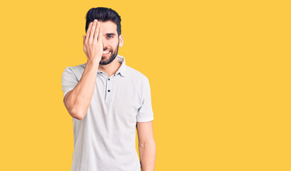 Young handsome man with beard wearing casual polo covering one eye with hand, confident smile on face and surprise emotion.