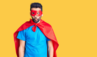 Young handsome man with beard wearing super hero costume making fish face with lips, crazy and...