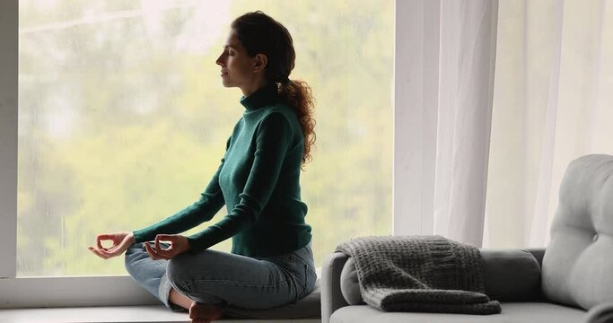 Serene woman in casual clothes sit cross-legged on pillow on windowsill with eyes closed do meditation, reducing fatigue, clear her mind, breath fresh air at home. Stress relief, yoga practice concept