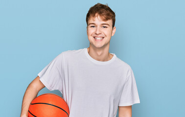 Obraz premium Young caucasian man holding basketball ball looking positive and happy standing and smiling with a confident smile showing teeth