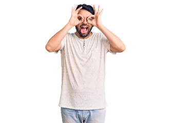 Fototapeta premium Young hispanic man wearing casual clothes doing ok gesture like binoculars sticking tongue out, eyes looking through fingers. crazy expression.