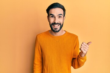 Young hispanic man wearing casual clothes smiling with happy face looking and pointing to the side with thumb up.