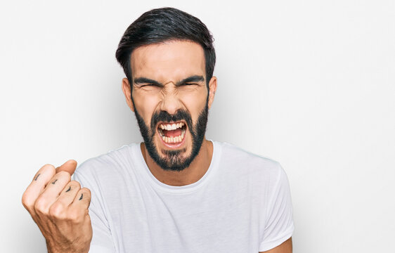 Young hispanic man wearing casual white t shirt angry and mad raising fist frustrated and furious while shouting with anger. rage and aggressive concept.