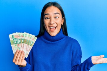 Young asian woman holding 50 hong kong dollars banknotes celebrating achievement with happy smile and winner expression with raised hand