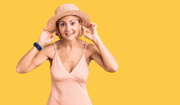 Young blonde woman wearing summer hat smiling pulling ears with fingers, funny gesture. audition problem