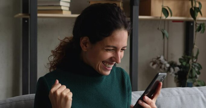 Woman sit on sofa holding smartphone read great news on sms feels overjoyed, clenched fist makes yes gesture looking very happy. Auction betting victory, personal achievement, candid emotions concept