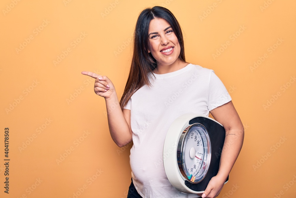 Poster young beautiful brunette pregnant woman holding controlling weight using weighting machine smiling h