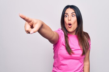 Young beautiful brunette woman wearing casual pink t-shirt standing over white background Pointing with finger surprised ahead, open mouth amazed expression, something on the front