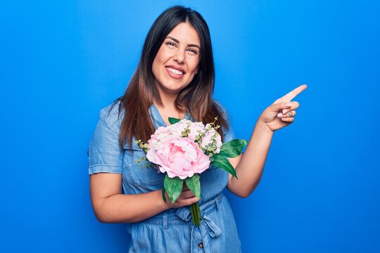 Young beautiful brunette woman holding bouquet of pink flowers over isolated blue background smiling happy pointing with hand and finger to the side