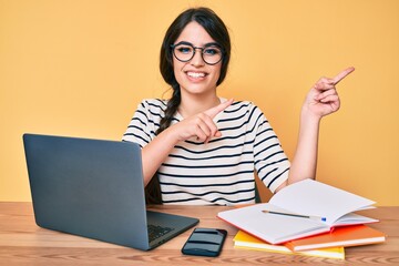 Brunette teenager girl working at the office with laptop smiling and looking at the camera pointing with two hands and fingers to the side.