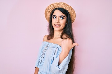 Brunette teenager girl wearing summer hat smiling with happy face looking and pointing to the side with thumb up.