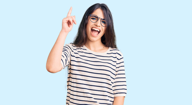 Brunette teenager girl wearing casual clothes and glasses pointing finger up with successful idea. exited and happy. number one.