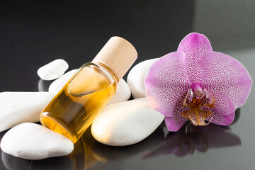 Lilac orchid flower and transparent glass bottle of yellow oil or perfume lying on white stones on a black surface