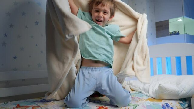 Slow motion of funny laughing toddler boy in pajamas hiding under blanket at night and jumping out. Scaring and frightening child.