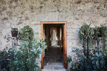 Fototapeta na wymiar Wedding dress hanging in a rustic room seen from outside the room in which you can see green plants