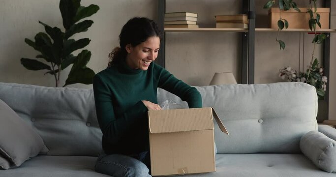 Happy young woman customer opening received parcel at home. Female shopper got purchased items, unbox box seated on couch feels satisfied by goods quality, condition and fast delivery services concept