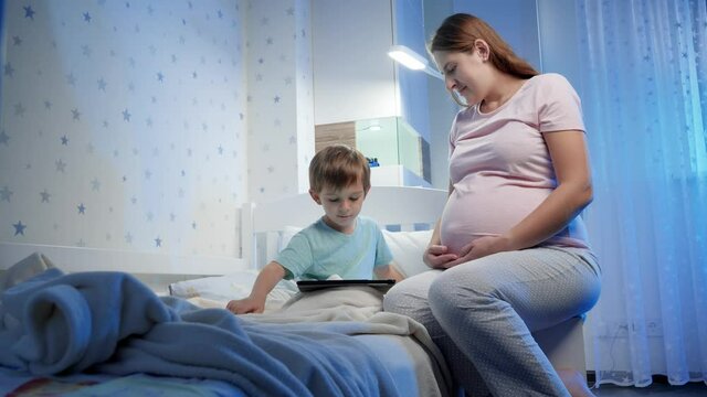 Young pregnant woman sitting at her little son bed and looking at him playing games on tablet computer. Children and adults using smartphones and gadgets at night during bedtime