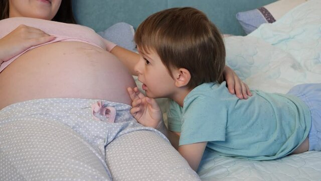 Portrait of little toddler boy in pajamas lying with pregnant mother in bed and talking to unborn baby in big belly. Loving and caring children