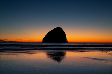 Fototapeta na wymiar A dramatic distant view of Haystack rock in silhouette, with a rich blue and golden sky at sunset, along the Pacific ocean coastline, Oregon