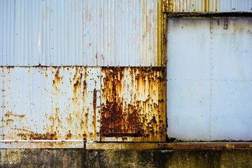 Part of the wall of a large building on a concrete base, sheathed with blue corrugated iron clapboard covered with rusty spots.