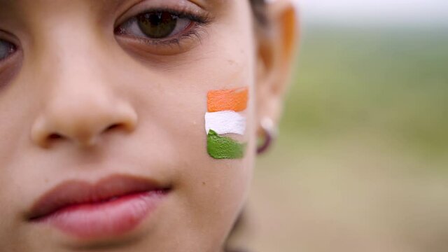 Close up selective focus of face, hands painting Indian flag tricolors on girl kid face during Independence or republic day celebration