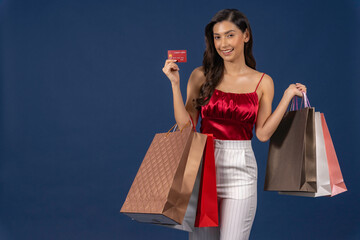 Young Asian woman holding credit card for online shopping on blue color background, black friday, cashless payment and consumerism concept, carrying shopping bags