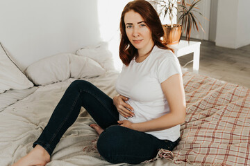 Fototapeta premium pregnant woman with red hair sits on the bed and holds her belly.