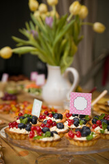Delicious homemade tartlets glazed with vanilla butter cream syrup and organic blueberries and red currants. Delicious sweet dessert for a party or celebration. A bouquet of tulips in the background
