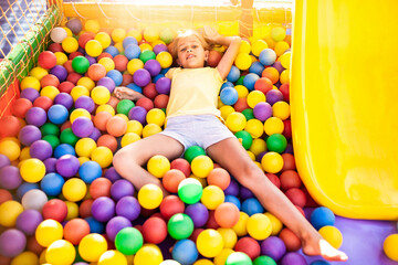 Fototapeta na wymiar A cute cheerful girl lies on the playground with soft and bright equipment and throws colorful balloons into the camera, enjoying the warm summer sun
