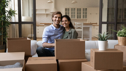 Portrait of smiling millennial couple renters unpack boxes in cozy living room in new home. Happy young Caucasian man and woman tenants feel excited unboxing relocate move to own house together.