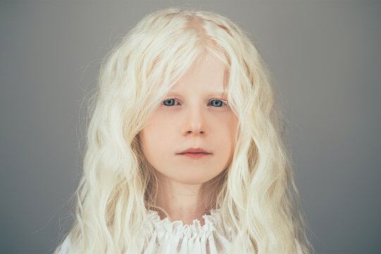 Beautiful little girl. Child innocence. Pure energy. Portrait of sweet peaceful albino blonde angel with curly hair blue eyes in white vintage blouse isolated on gray background.