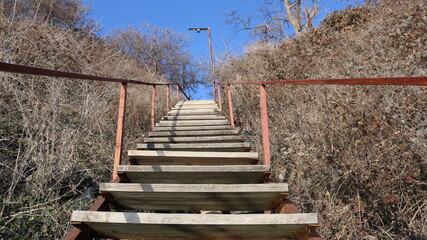 wooden staircase with metal railings visible from the bottom up, going high up the hill overgrown with dry grass and bushes to the lantern against the background of a bright blue sky, retro element