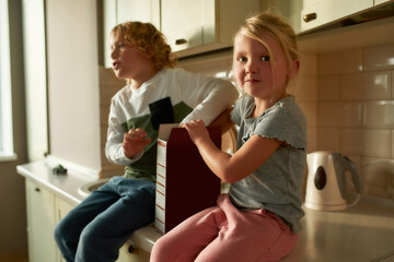 Portrait of cute little girl looking at camera, eating cornflakes while sitting on the kitchen cabinet together with her brother at home