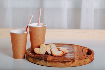 Heart-shaped cookies on round wooden cutting board. Saint Valentine's cookies in shape of heart and craft coffee cups on white background. Two paper coffee to go with tubes.