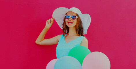 Happy smiling young woman with colorful balloons wearing a summer straw hat on a pink background