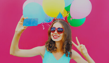 Portrait of cheerful smiling woman taking selfie picture by smartphone with colorful balloons on a pink background