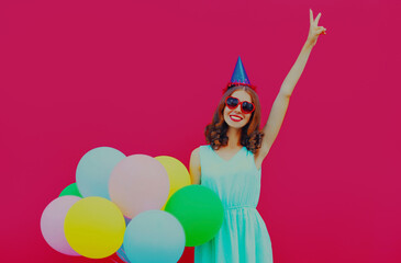 Obraz na płótnie Canvas Happy surprised smiling young woman in birthday cap with colorful balloons on a pink background