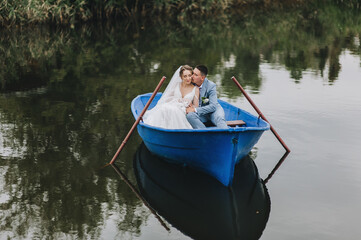 Stylish groom in a suit and a beautiful bride in a white lace dress are sitting in a wooden boat and swimming on the lake, enjoying nature.