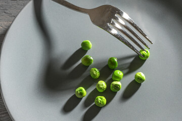 Some green peas and a fork with shadows on a gray plate, meager diet meal after the resolution to...