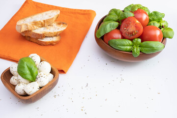 fresh tomatoes, mozzarella and green basil in a wooden bowl, near croutons and spices on a white table