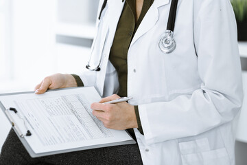 Unknown woman-doctor writing something at clipboard while sitting at the chair, close-up. Therapist wearing green blouse at work is filling up medication history record. Medicine concept