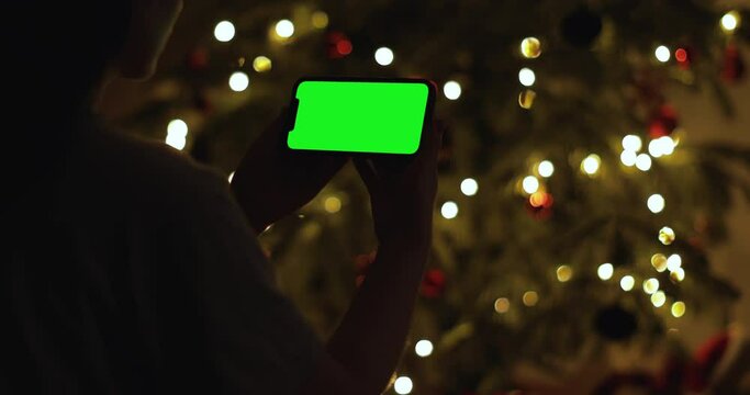 Brunette woman is sitting on a dark room and uses a mobile phone with Green Screen for Compositing in front of the Christmas tree. New Year Celebration Concept. 