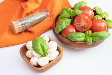 fresh tomatoes and green basil in a wooden bowl, next to mozzarella in a wooden bowl and spices on a white table